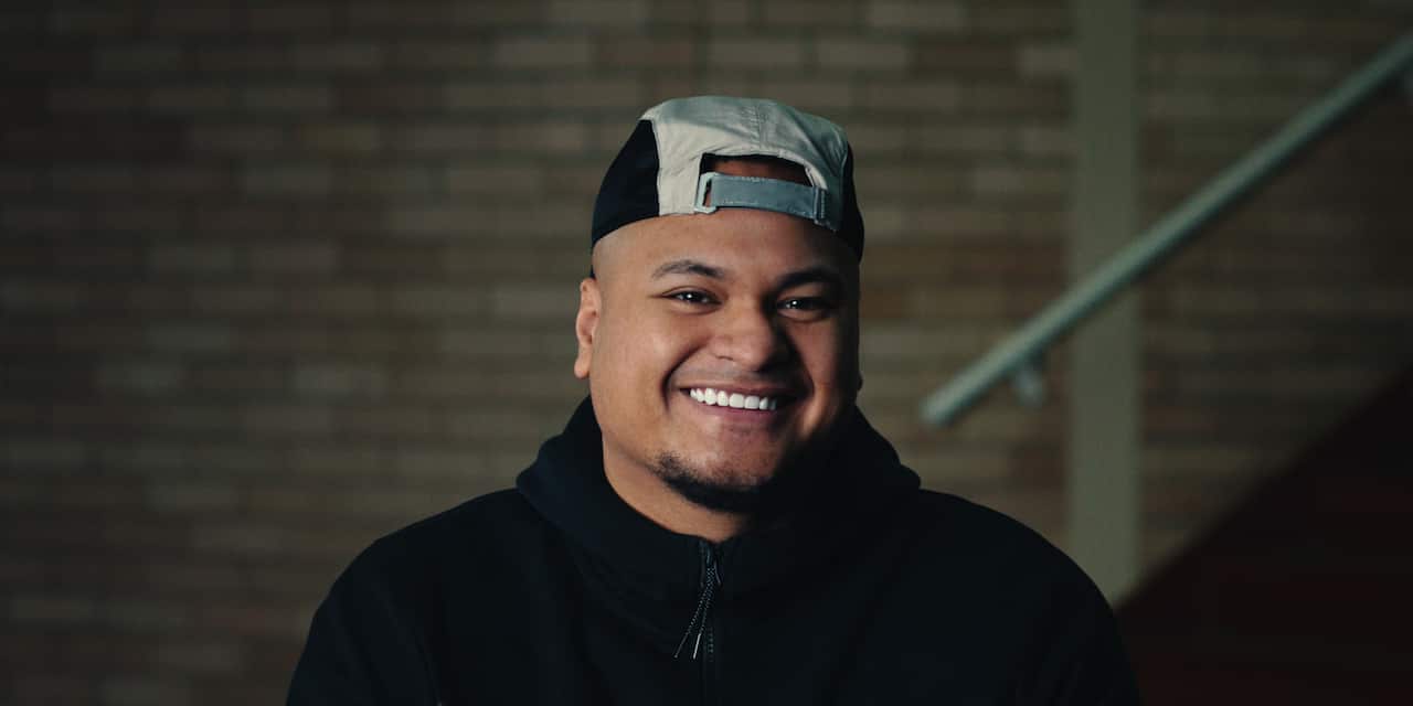 A man in a black hoodie smiling in front of a brick wall.