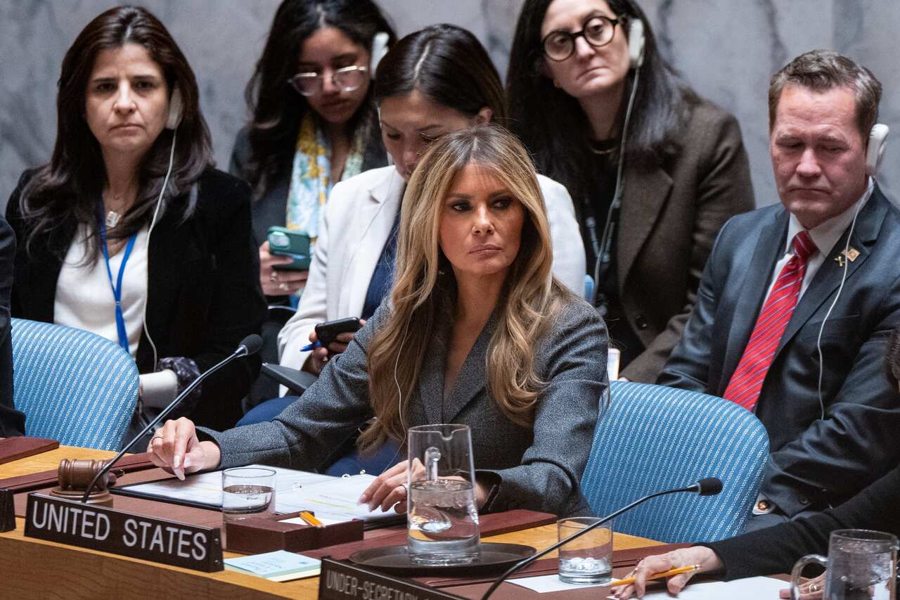 A woman with long hair in a suit sits in an official meeting room among other people behind a sign that reads 'United States'.