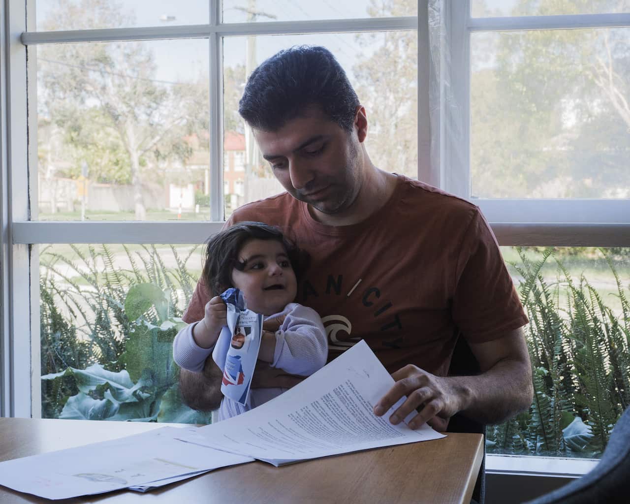 A man sits at a table near a large window, holding a young child in his lap while looking down at a stack of papers. The child is scrunching a sheet of paper and looking towards the side.