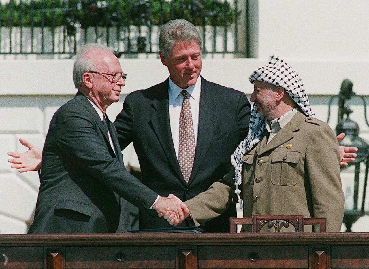 Two men shake hands in front of a white building. A man stands between them with his arms outstretched around them. The man in the middle and the man on the left wear dark suits. The man on the right wears a military jacket and a black and white keffiyeh on his head.
