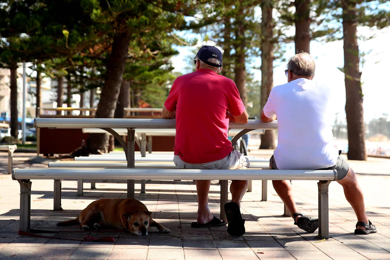 Two older men, their backs turned, sit at a picnic table while a dog rests at their feet.
