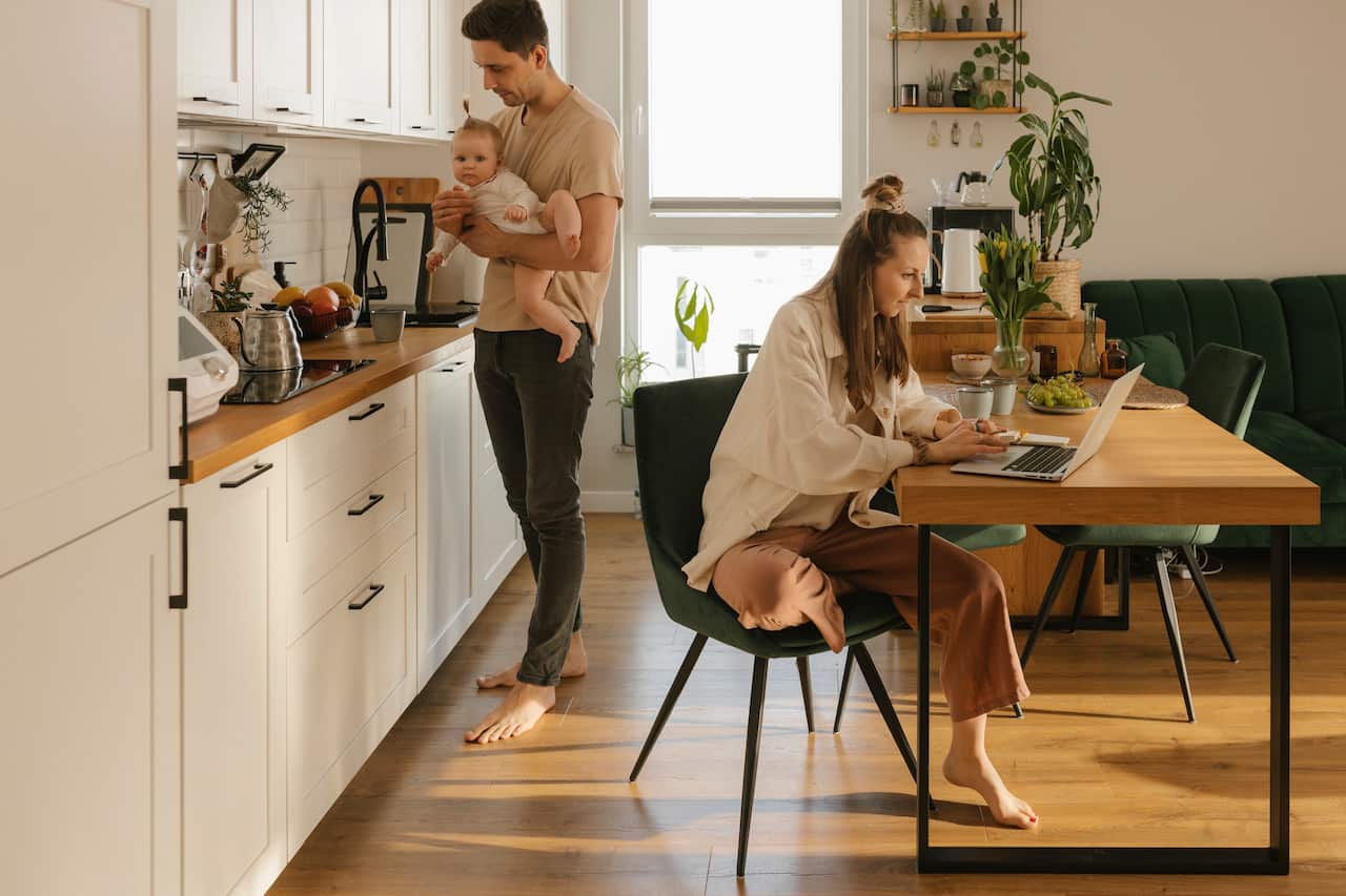 A woman works on a laptop in the kitchen area while a man carrying a baby walks by.