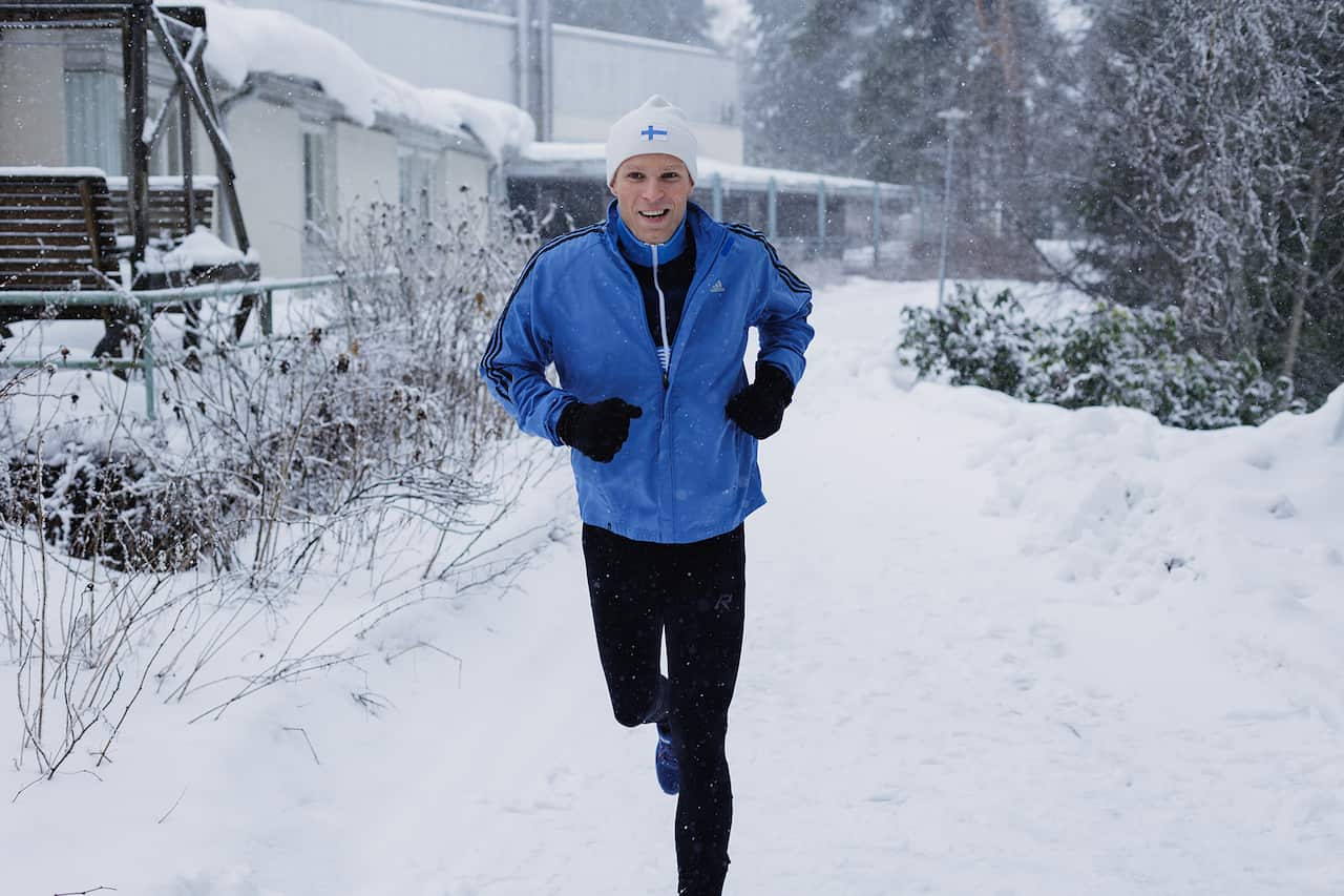 A man in black pants, blue jacket and white beanie runs along a snowy road, beside a long building.