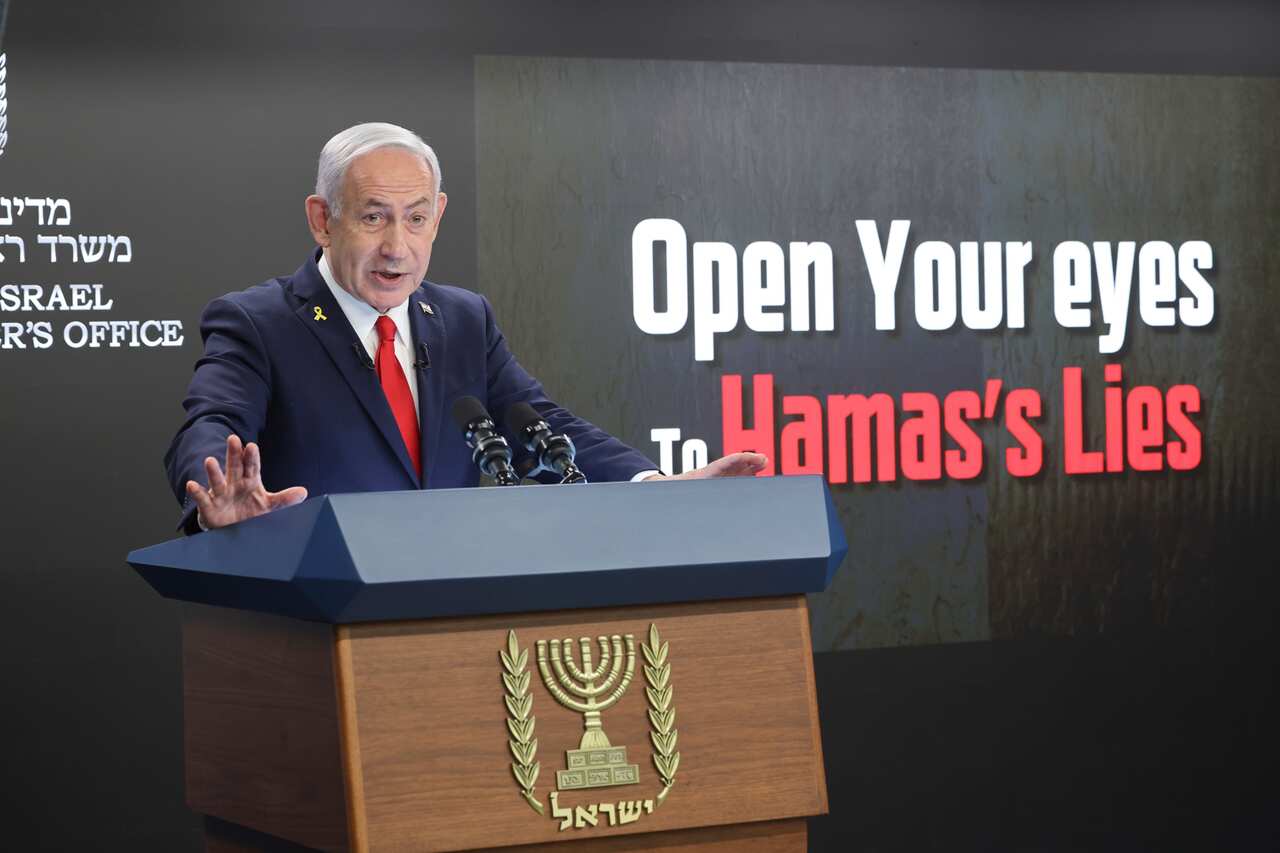 A man in a dark suit and a red tie speaks as he stands at a lectern.