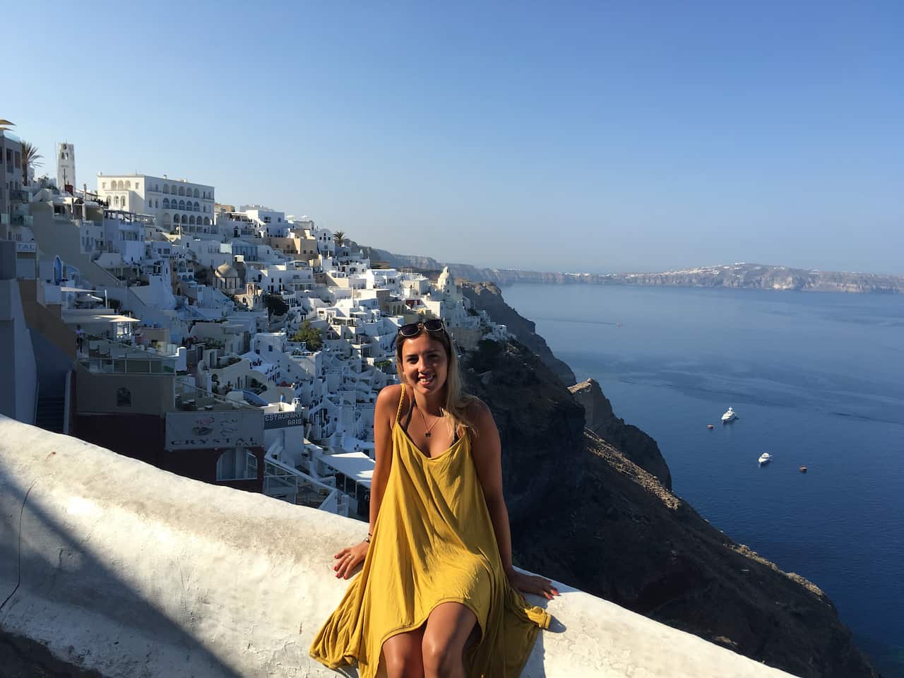 A woman in a yellow summer dress sits on a wall. In the background there's a whitewashed Greek town and the Aegean Sea.