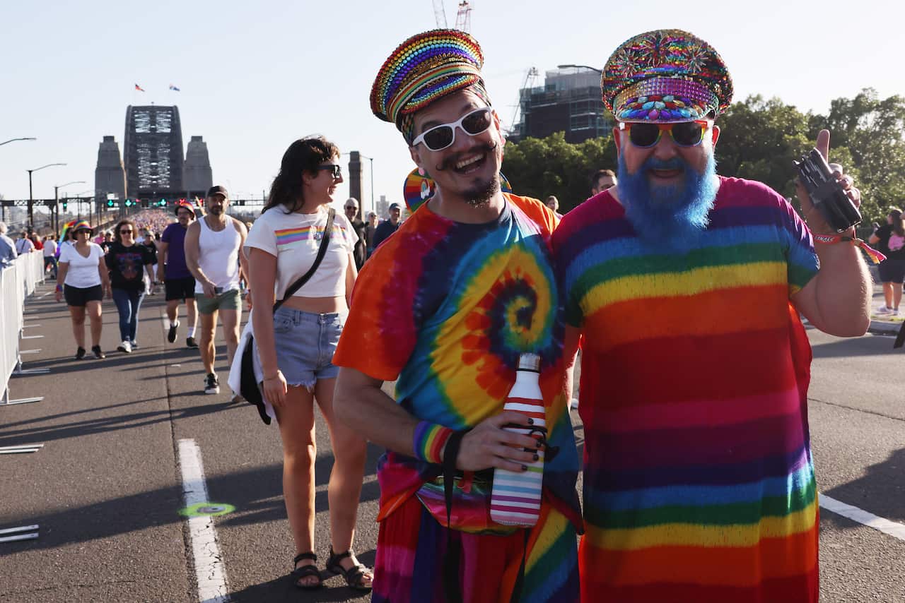 Two men in rainbow-coloured tops and hats pose with the Sydney Harbour Bridge in the background