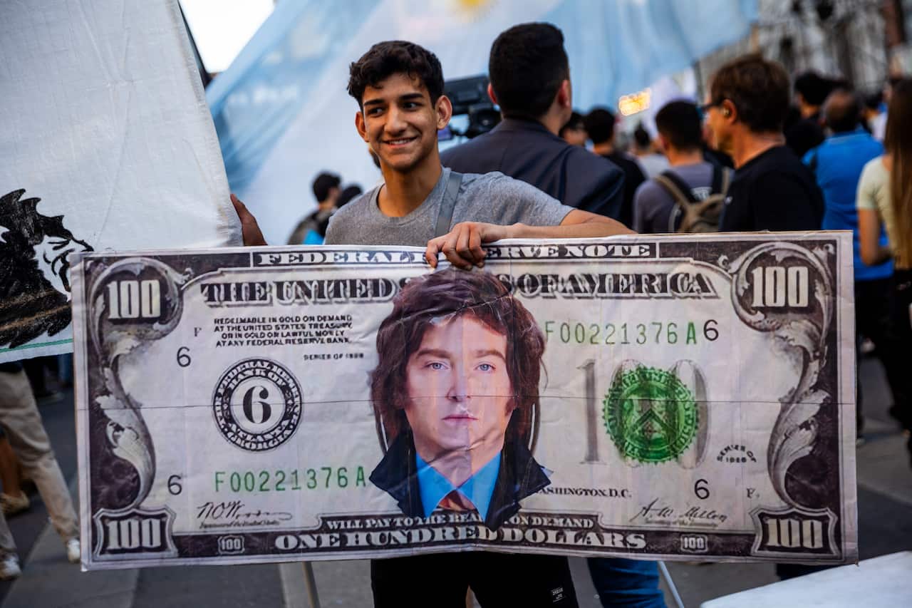 A man holding up an oversized US $100 bill with the face of Javier Milei on it.