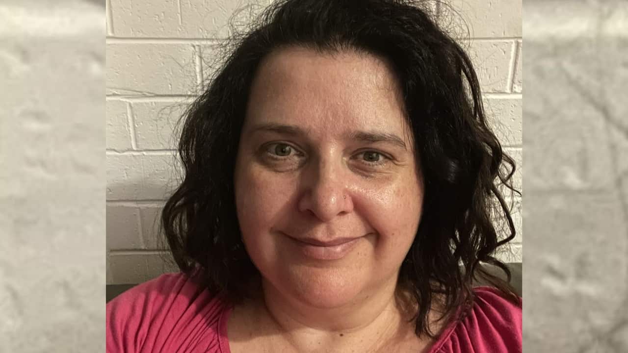 a woman with dark hair worn out in a pink shirt smiles in a close up pic towards camera