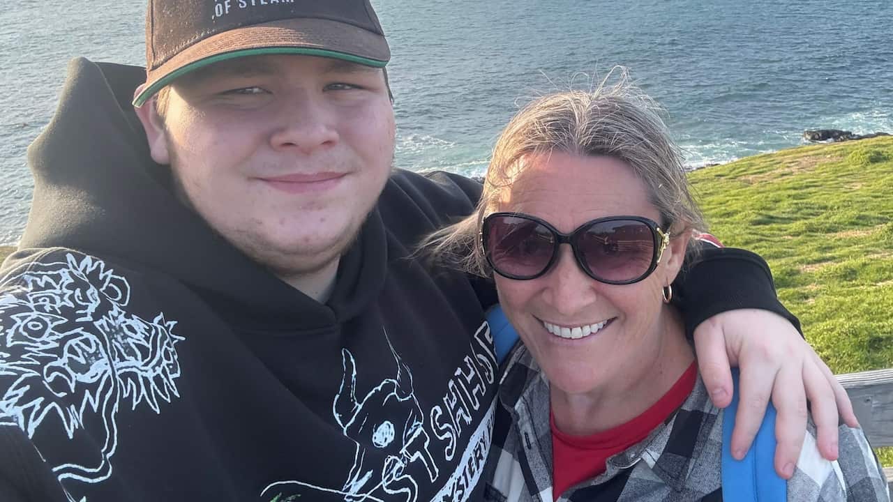 a teenage boy has his arm around his mum - they stand on a grassy headland