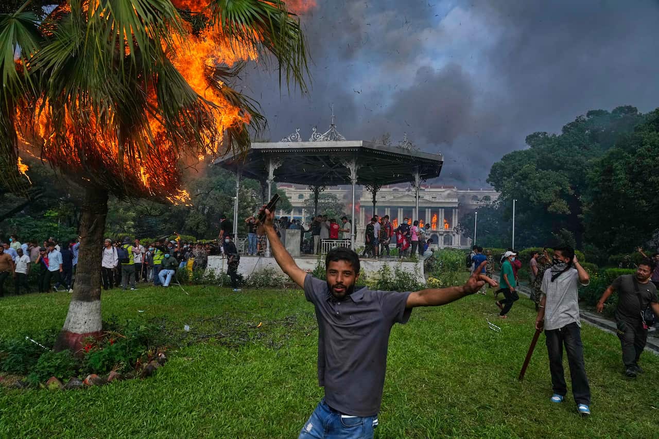 A man stands with his arms outstretched in the foreground of a park, with a crowd of people gathered behind him. In the background, a building is visible with flames coming from its windows, and a palm tree in the left of the frame is also on fire.