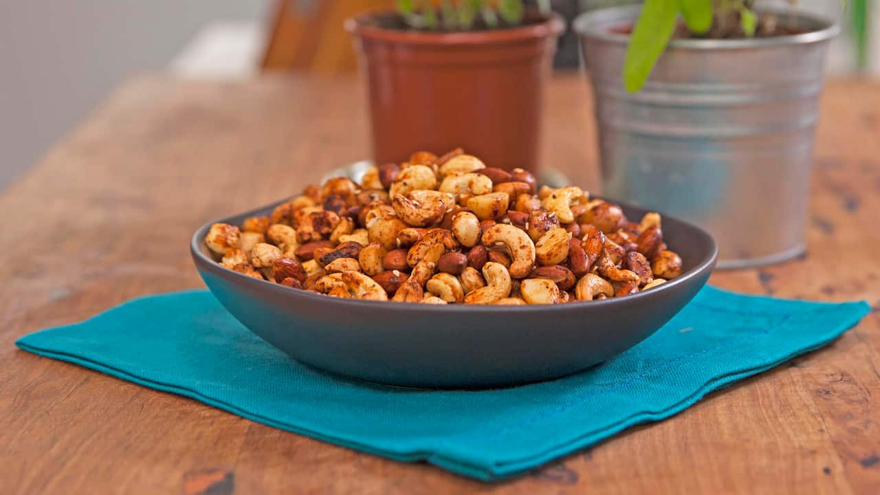 A dark grey bowl,  piled with cooked mixed nuts, sits on a blue napkin on a wooden surface. 