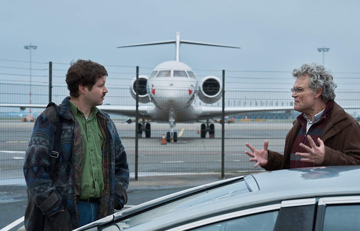 Two men stand beside a car, one talking animatedly. A plane on a runway can be seen in the background.