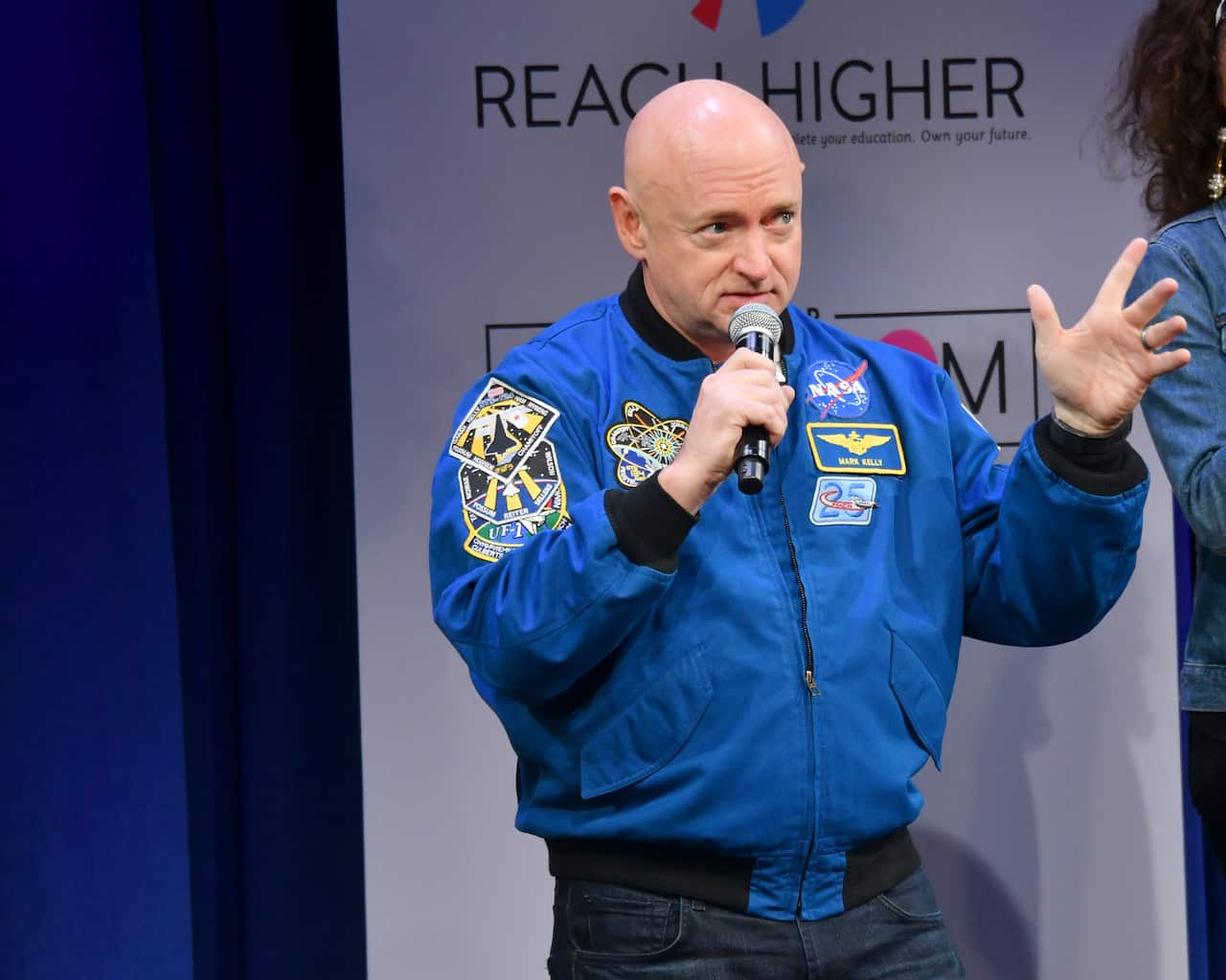 A bald man talking into a microphone, with a blue jacket that has NASA and navy officer stickers.
