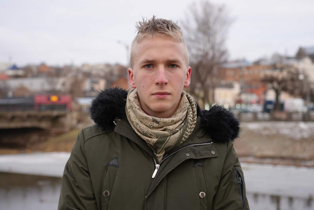 A 19-year old man with blond hair wearing a jacket and scarf and standing outside.