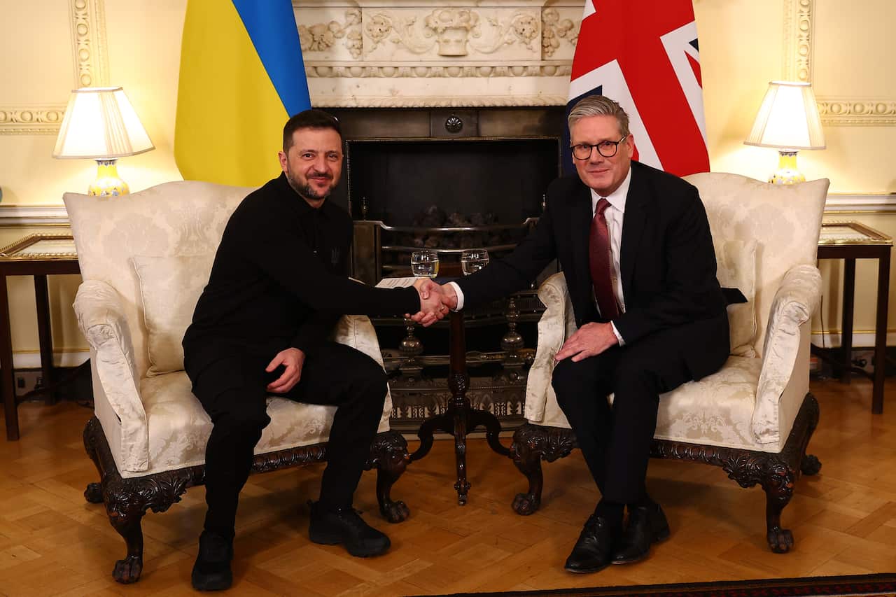 Ukrainian President Volodymyr Zelenskyy shakes UK Prime Minister Keir Starmer's hand. They are sitting on cream-coloured arm chairs and the flags of their respective countries are behind them