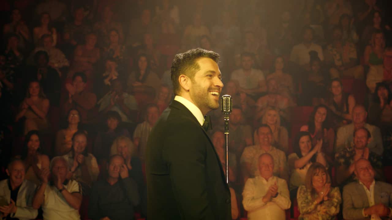 A smiling man stands side on in the image, near a tall microphone. A gentle spotlight illuminates his face. Rows of audience members can be seen behind him, applauding.