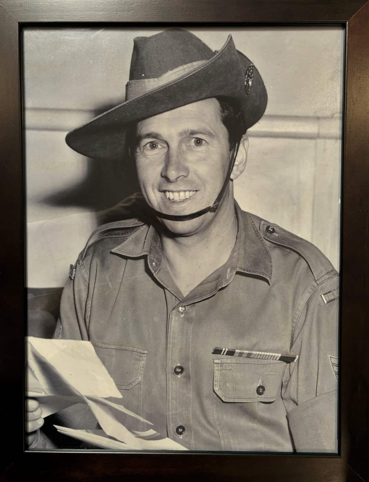 An old photo of an Australian soldier in a uniform with a hat and holding up a document is smiling.