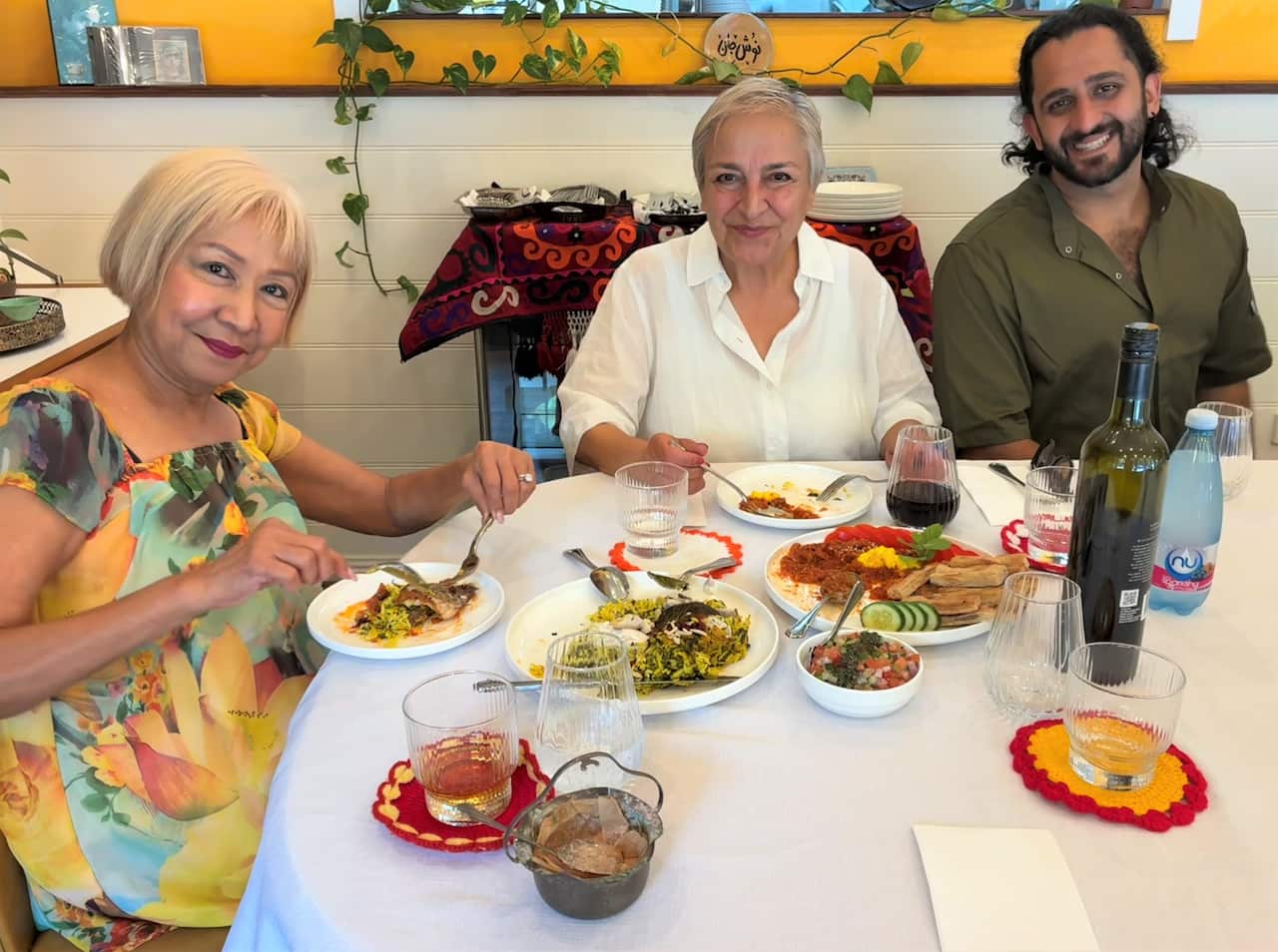 Two women sit at a restaurant table laden with food, next to a man with a beard smiing at camera.