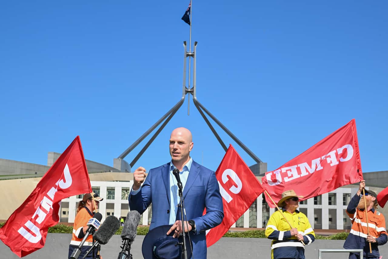 Man in a suit holding a hat speaks at a microphone in front of Parliament.
