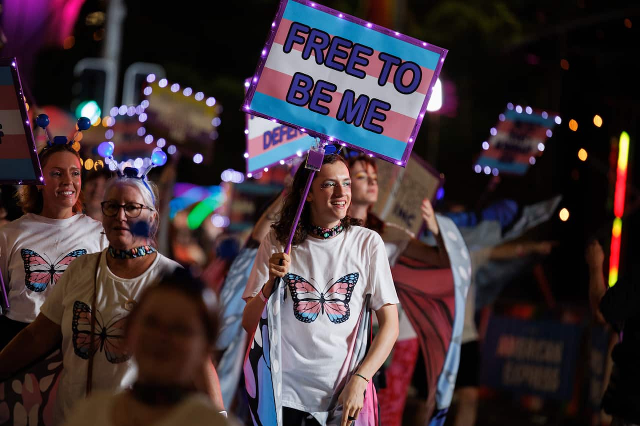 A young person wearing a t-shirt with a butterfly in the colours of the trans flag on it. They are holding a sign that says "free to be me".
