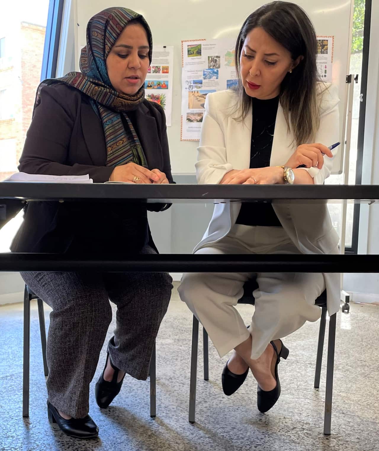 A woman in a brown jacket sits at a desk with another woman.