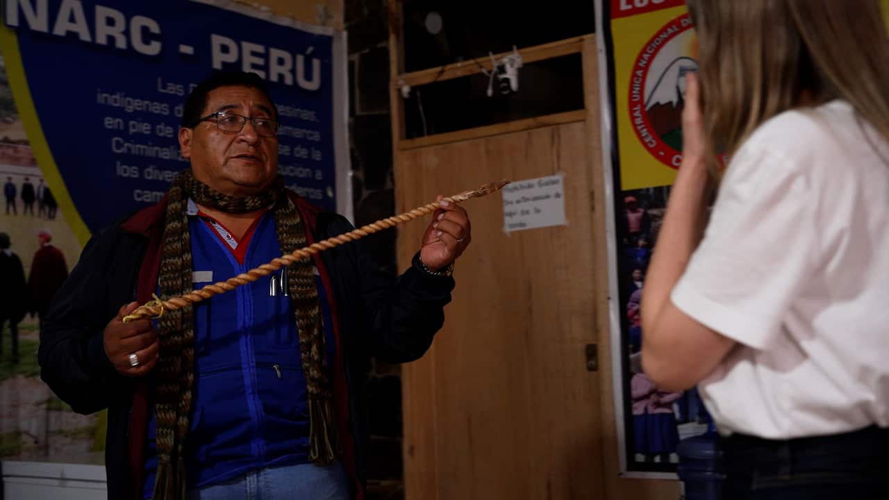 A man holds a long, braided stick between two hands. He is showing it to a woman who is standing in the corner of the frame, her face obscured. 