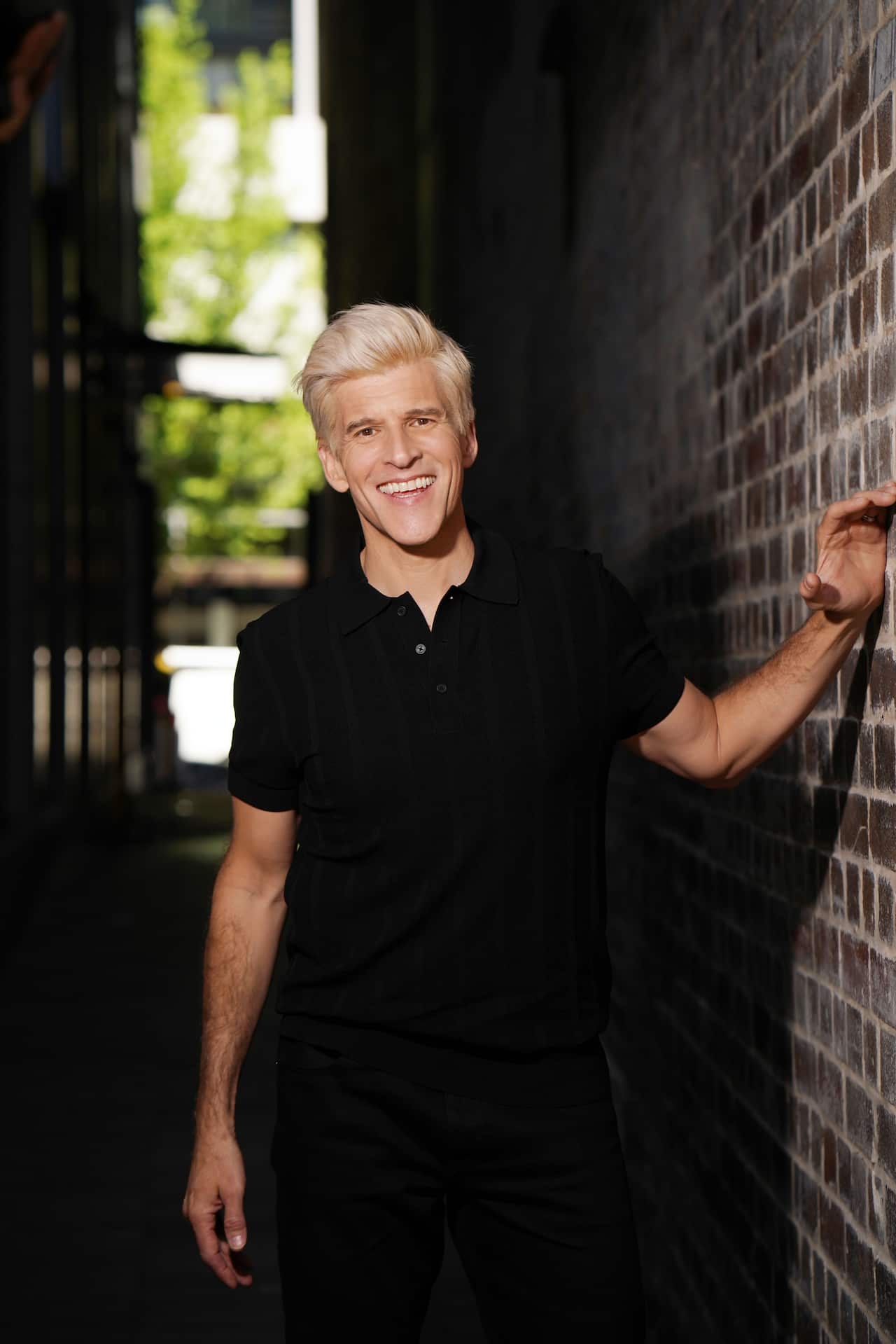 A man stands outside, hand on a brick wall and smiles.