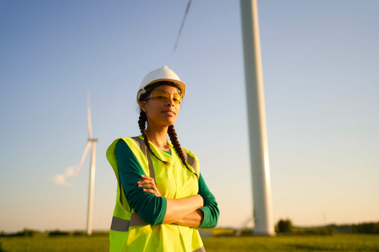 Woman wears high-vis vest and stands with her arms folded in front of a wind turbine. She's wearing a white hard hat, green long sleeve jumper under the vest and has two braids.