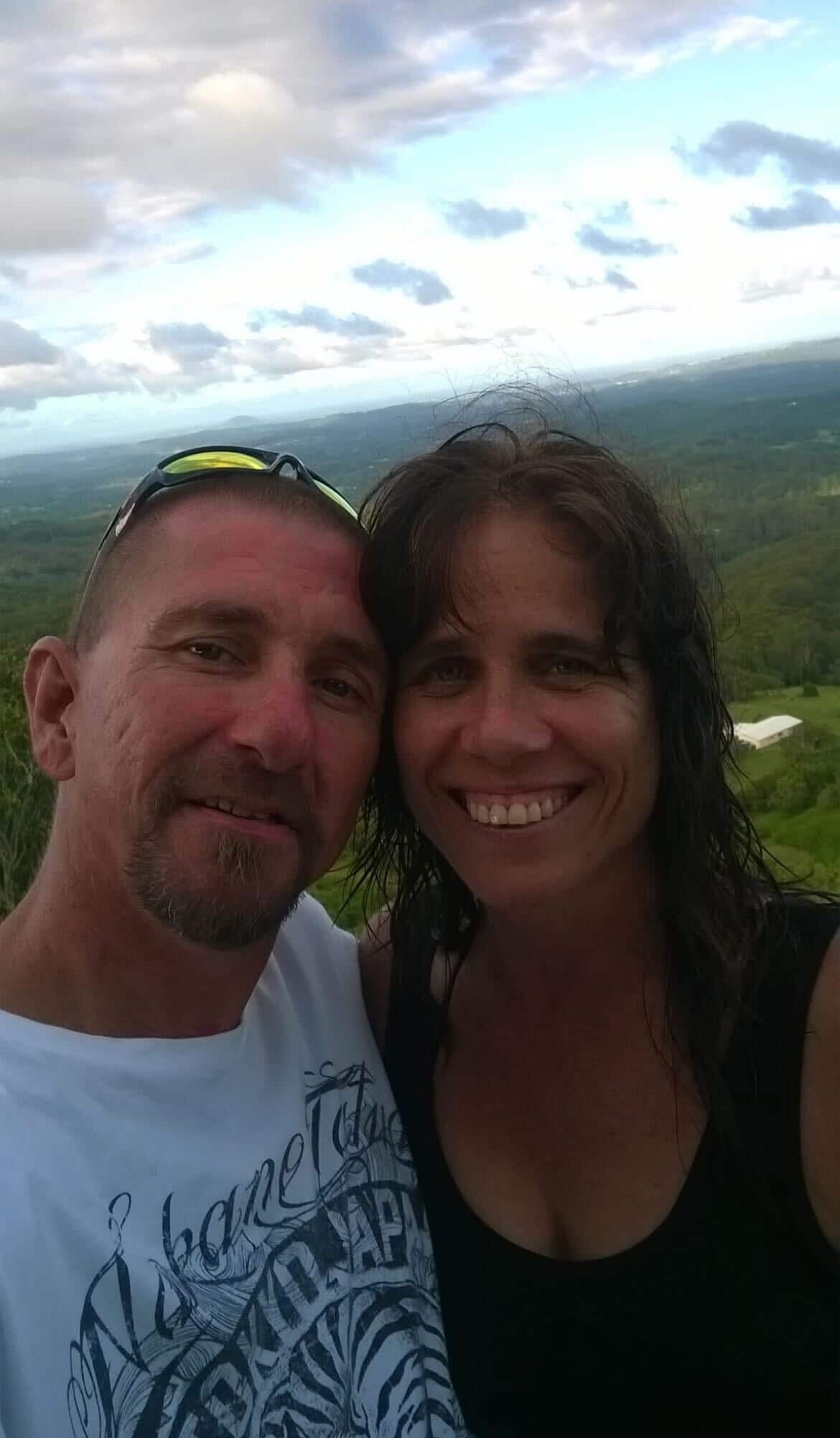 A man in a white graphic T-shirt with sunglasses on his head stands next to a smiling woman in a black top with brown hair