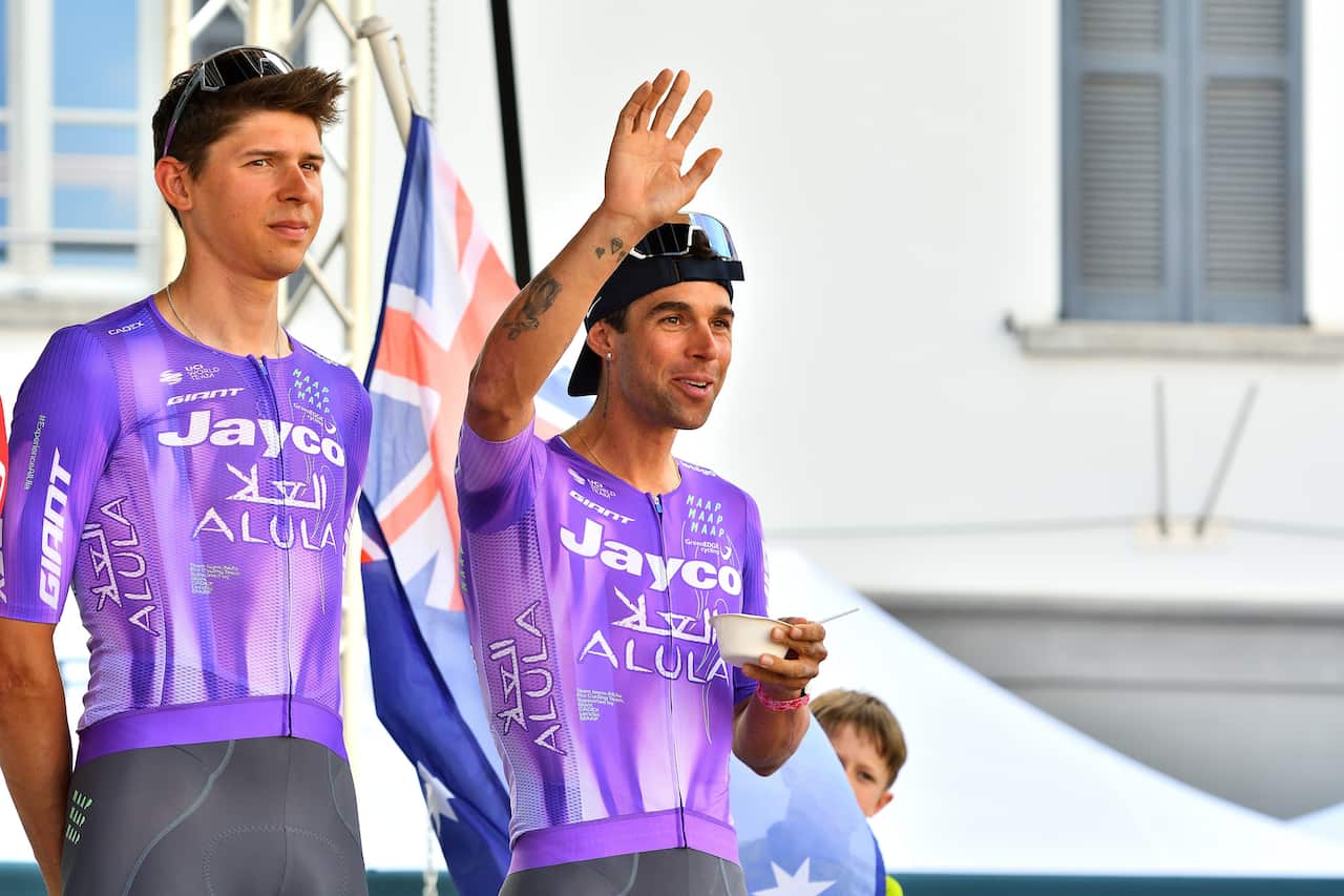 Two men in purple cycling jerseys standing together. One is raising his right hand