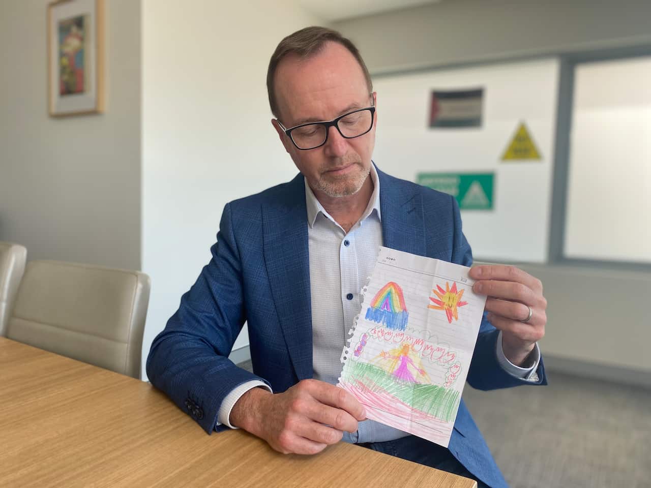 A bespectacled man in a blue, textured blazer and white shirt sits at a wooden table, looking down at a child's crayon drawing he holds in both hands. The drawing, on a piece of lined paper, features a brightly coloured rainbow, a smiling sun, and a central figure in a pink dress surrounded by colourful, flame-like shapes.