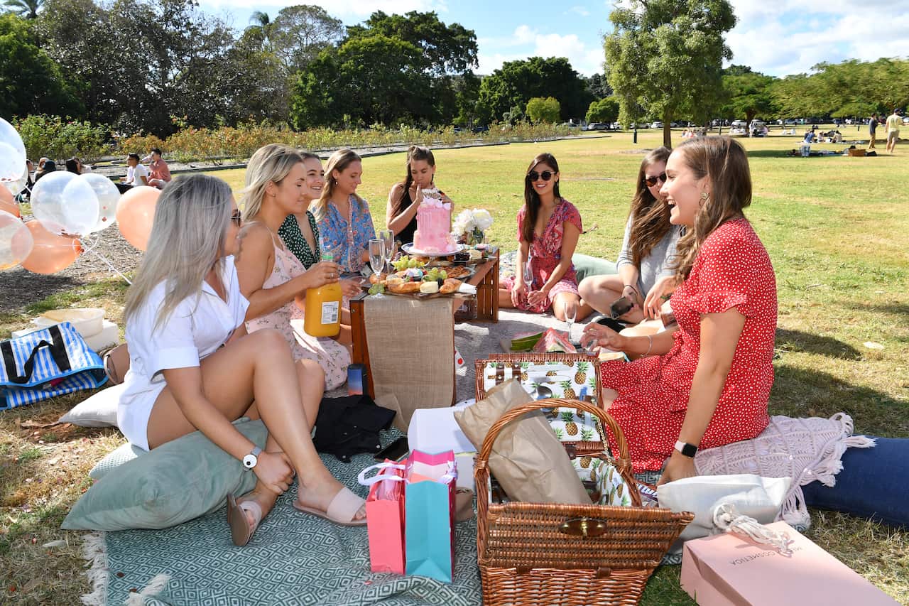 A group of young women sit on picnic rugs on grass. They are seen smiling and talking to one another.