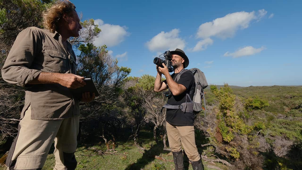 Standing in the outdoors, a man with a camera films another man.