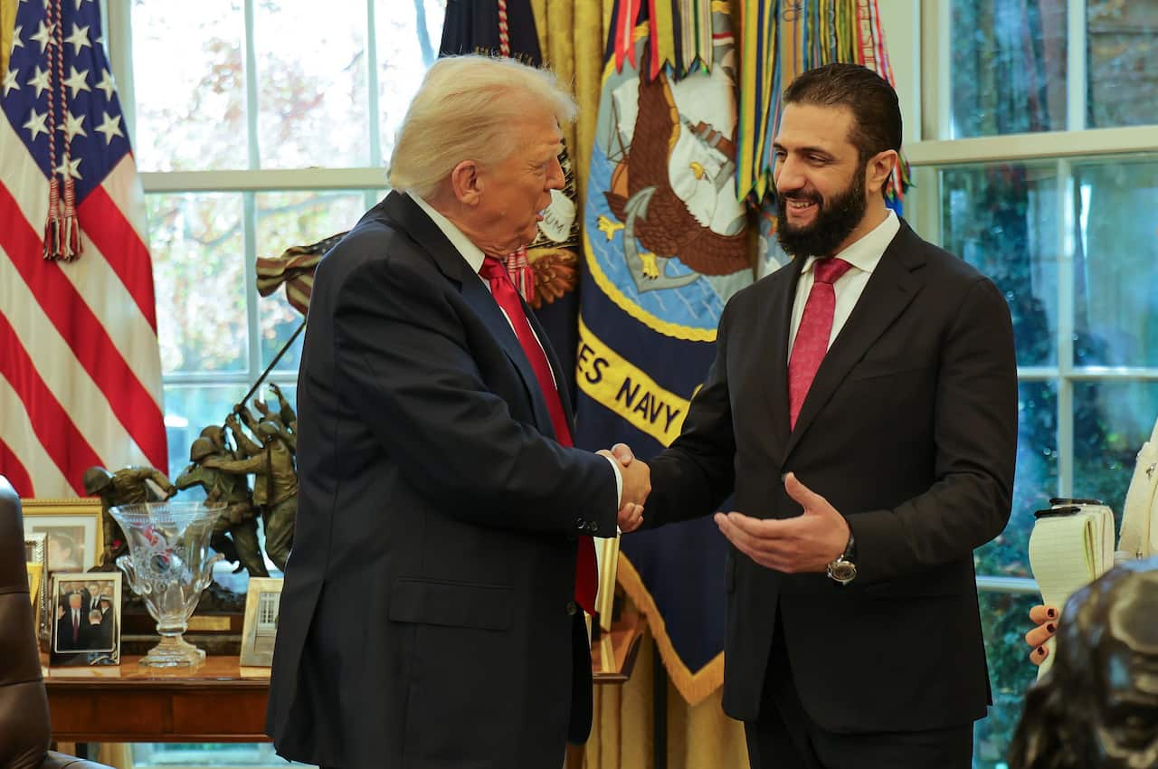 Donald Trump and Ahmed al-Sharaa shake hands in the Oval Office. They are both wearing suits, and appear to be mid-conversation. 