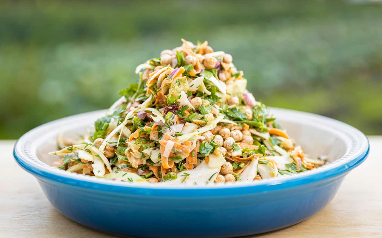A pile of colourful cabbage, herb and chickpea salad sits in a bright blue serving dish on a wooden table. 