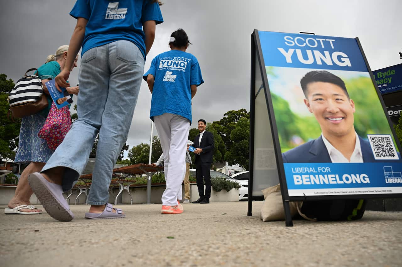 A blue Liberal corflute is positioned on the ground, as volunteers hand out Liberal how to vote cards.