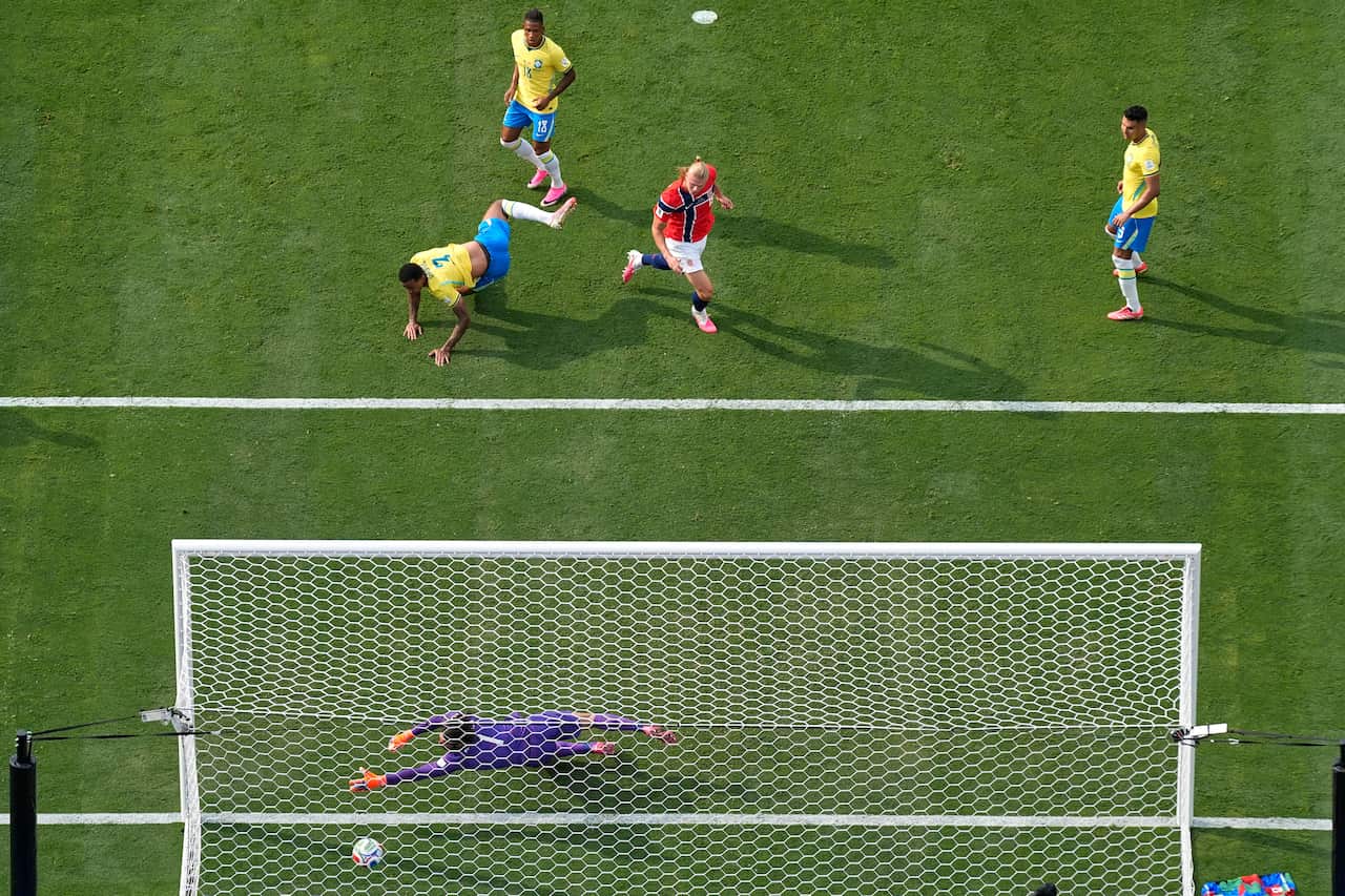 A goalkeeper wearing purple lunges for a football, as other players crowd around the goal net.