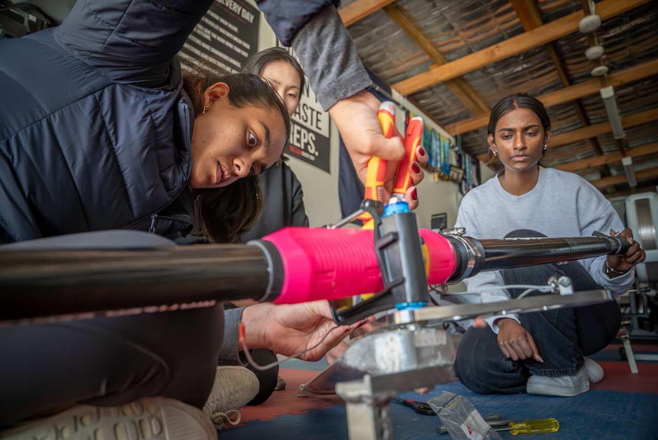 Three girls fix a device attached to an oar. One holds pliers. 
