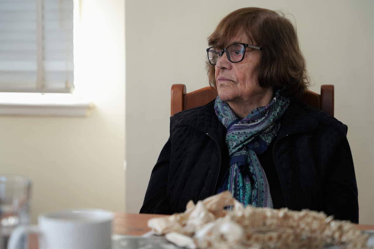 An older woman with short brown hair sits in a wooden chair, wearing a black jacket with a patterned blue scarf and black-rimmed glasses, looking off to the side.