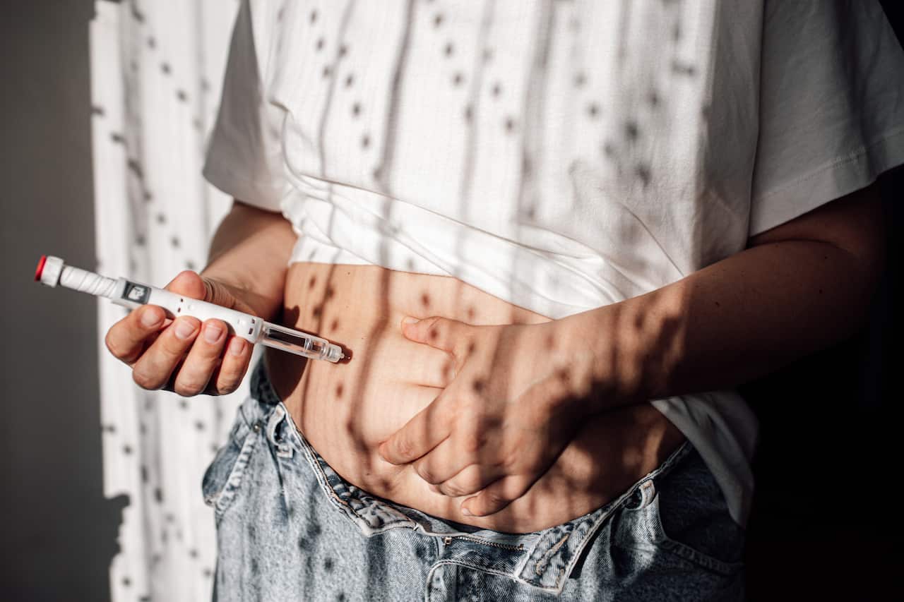 A person wearing blue jeans and a white shirt holding a syringe against their exposed stomach