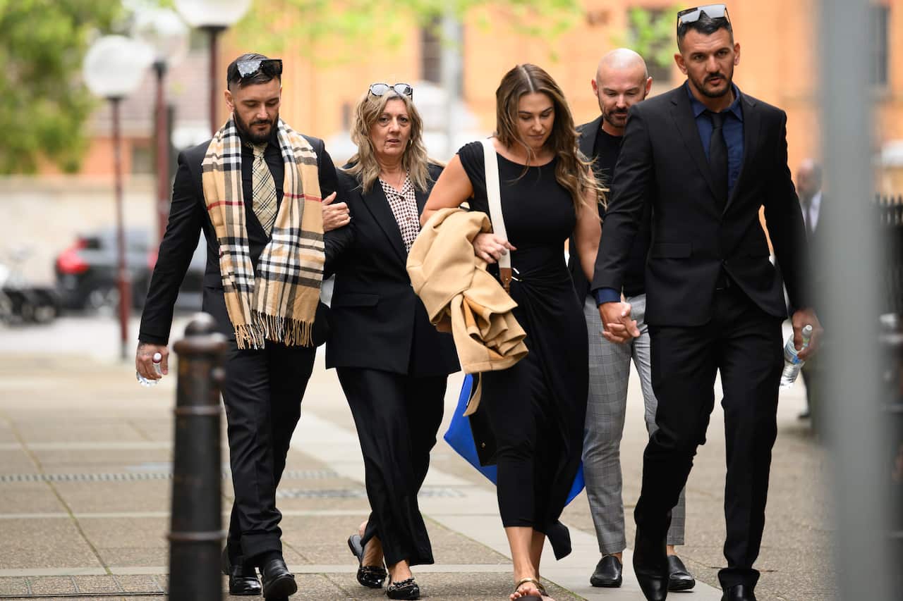 A group of five people dressed in dark formal clothing walk together along a city footpath outside a court building.