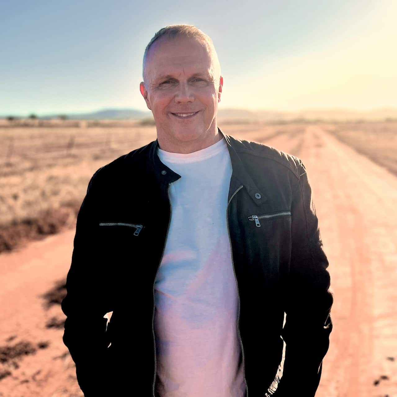 A man in a black jacket and white t-shirt smiling while standing on a dirt road