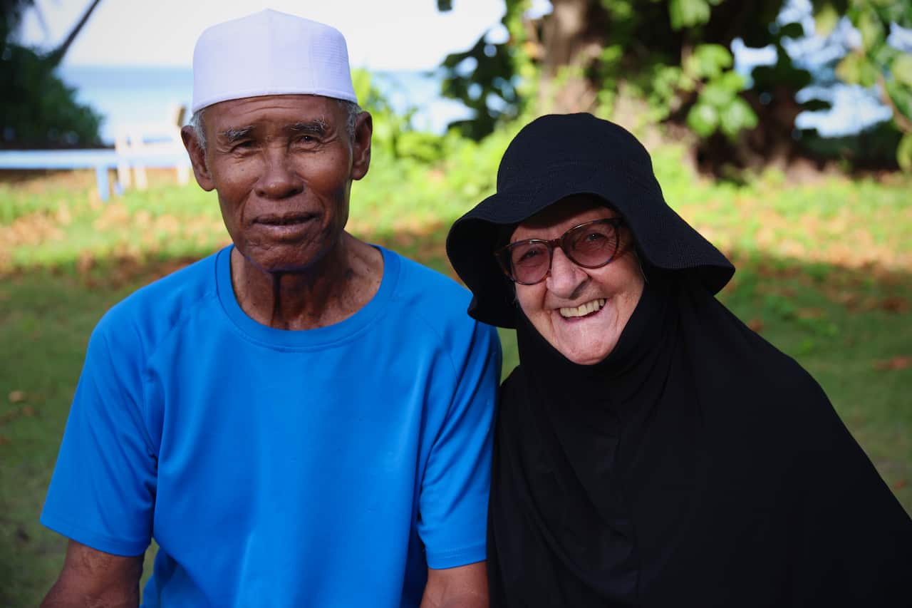 A man wearing a white cap and blue T-shirt sits next to a woman in a burqa in a garden. Both are smiling.