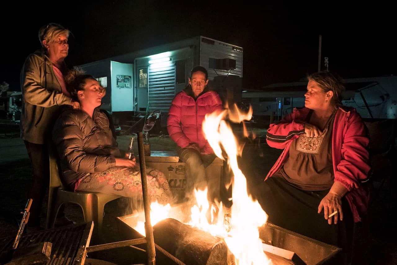 Four women sit outdoors, around a wood fire. A mobile home can be seen behind them.  