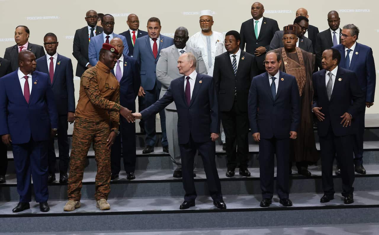 A group of politicians in suits pose for a group photo while a man in the middle is shaking hands with a young man in military fatigues and red beret
