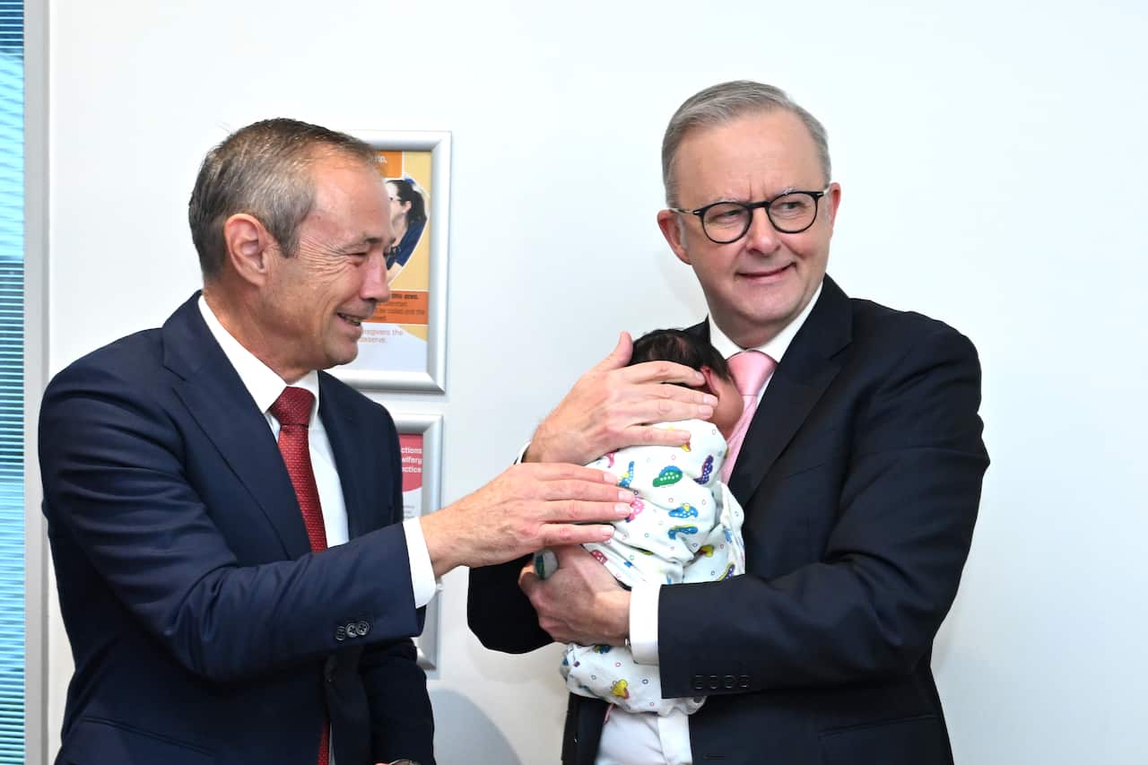 Anthony Albanese, dressed in a suit, embraces a baby while another man in a suit places his hand on the baby, looking at them both.