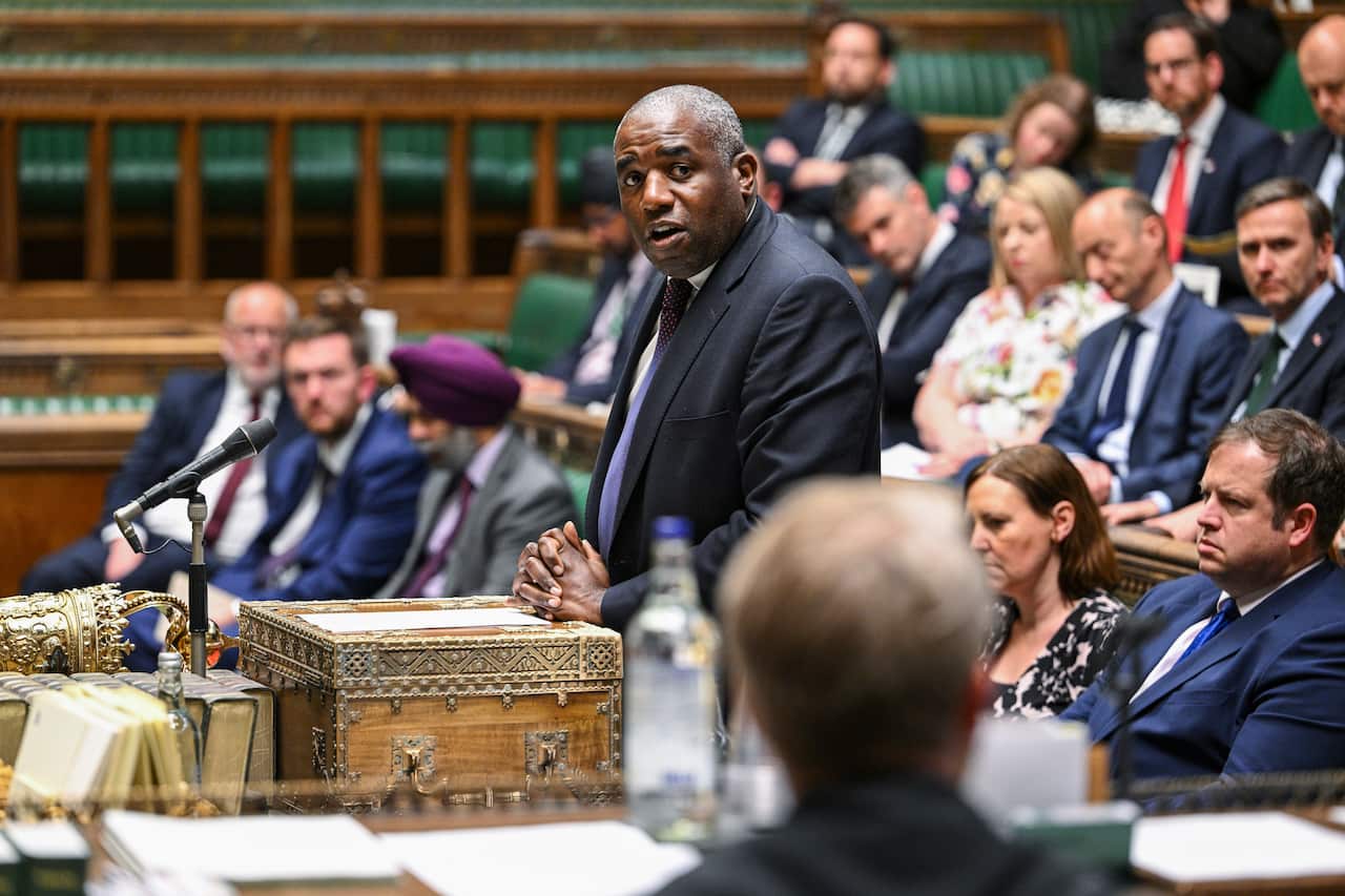 A man standing up at a microphone on top of an ornate box, in a parliamentary chamber as people sit on benches around