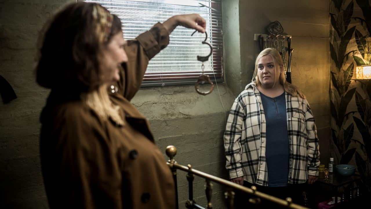 A young woman looks at an older woman holding up a set of handcuffs. They are standing in a room of a brothel.jpeg