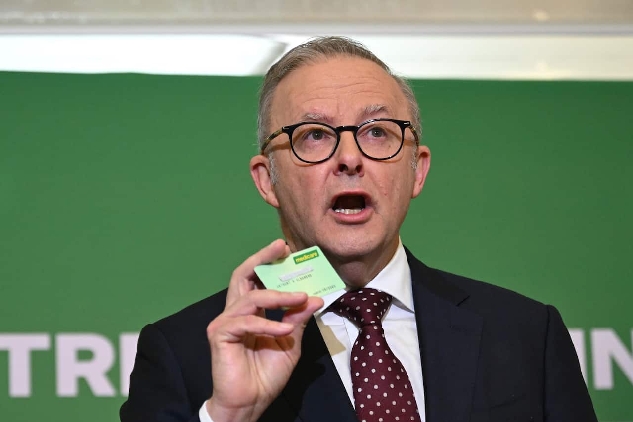 A man in a suit and tie is holding up a small card while speaking. He is wearing glasses and has gray hair. A green backdrop is visible behind him.