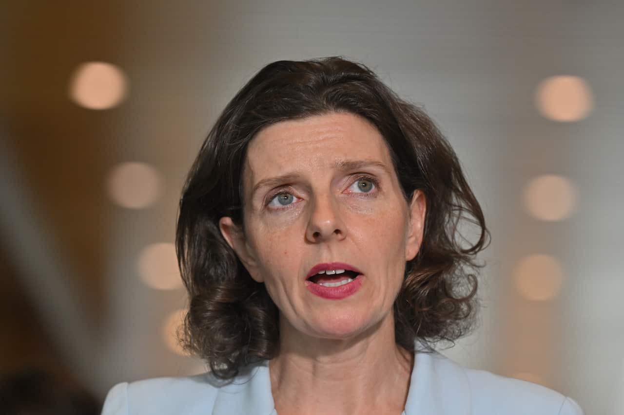 A woman with brown hair and a blue shirt speaking in a hallway.